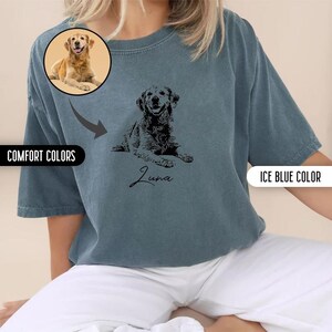 K&ouml;nnte beinhalten: Ein blaues T-Shirt mit einer schwarz-wei&szlig;en Illustration eines Hundes und dem Namen "Luna" in Kursivschrift. Das Shirt wird von einem Model getragen, das eine wei&szlig;e Hose tr&auml;gt.