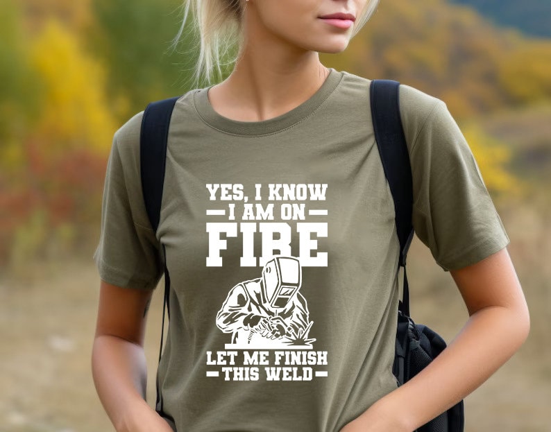 May include: A green t-shirt with a white graphic of a welder with the text "YES, I KNOW - I AM ON FIRE - LET ME FINISH - THIS WELD - "