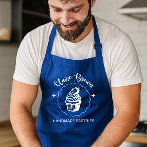 May include: A baker wearing a blue apron with the text "Claire Brown Handmade Pastries" and a cupcake design. The baker is working with dough on a wooden surface, with flour dusted on the surface and the baker's face.
