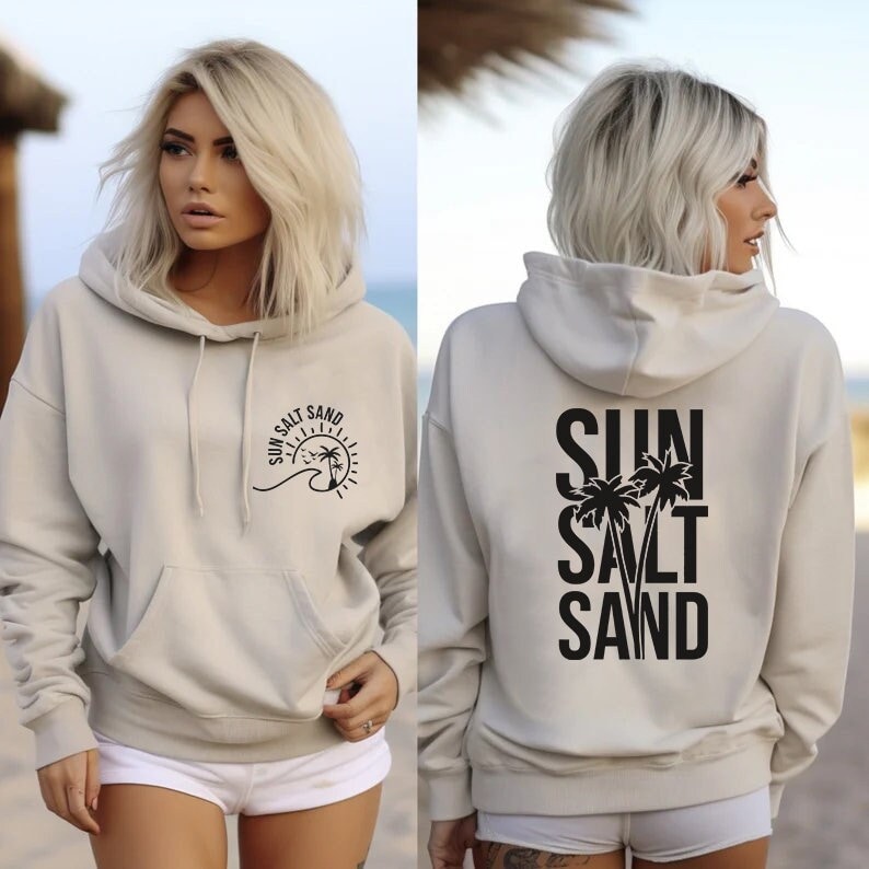 May include: A light beige hoodie with a black graphic on the front that says "Sun Salt Sand" with a sun, wave, and palm trees. The back of the hoodie has a larger black graphic that says "Sun Salt Sand" with palm trees.
