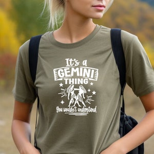 May include: Olive green t-shirt with the text "It's a GEMINI thing, you wouldn't understand" in white. The graphic includes two figures and stars. The shirt is a short-sleeved crew neck.