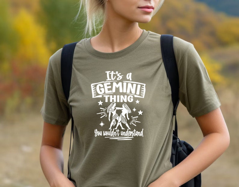 May include: Olive green t-shirt with the text "It's a GEMINI thing, you wouldn't understand" in white. The graphic includes two figures and stars. The shirt is a short-sleeved crew neck.