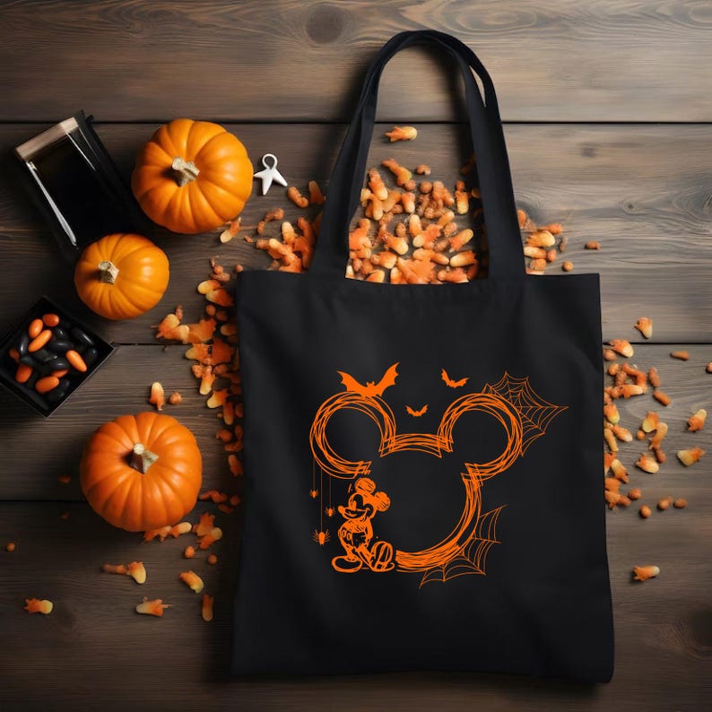 Puede incluir: Una bolsa de tela negra con un gr&aacute;fico naranja de una silueta de Mickey Mouse delineada en estilo garabateado. El dise&ntilde;o incluye peque&ntilde;os murci&eacute;lagos, telara&ntilde;as y una peque&ntilde;a figura de Mickey Mouse de pie en la base del contorno circular.