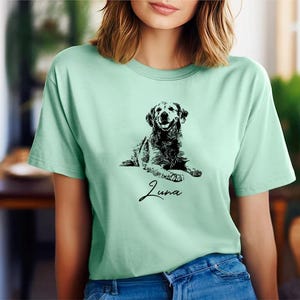 May include: A light green t-shirt featuring a black and white illustration of a dog. The dog is lying down and the text "Luna" is printed below the dog.