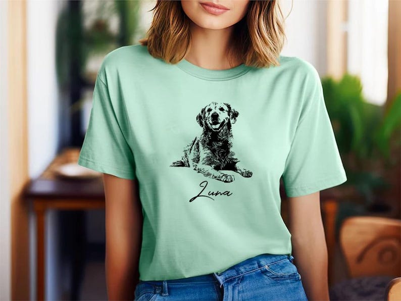 May include: A light green t-shirt featuring a black and white illustration of a dog. The dog is lying down and the text "Luna" is printed below the dog.
