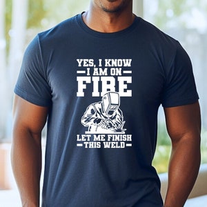 May include: A navy blue t-shirt with white text that reads "Yes, I know - I am on - FIRE. Let me finish - this weld -." The shirt features a white silhouette of a welder wearing a welding mask.