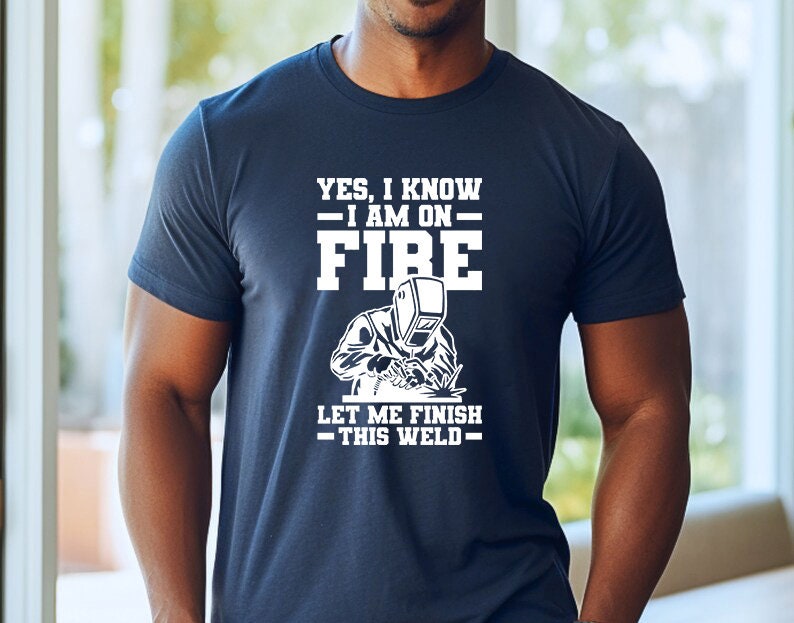 May include: A navy blue t-shirt with white text that reads "Yes, I know - I am on - FIRE. Let me finish - this weld -." The shirt features a white silhouette of a welder wearing a welding mask.
