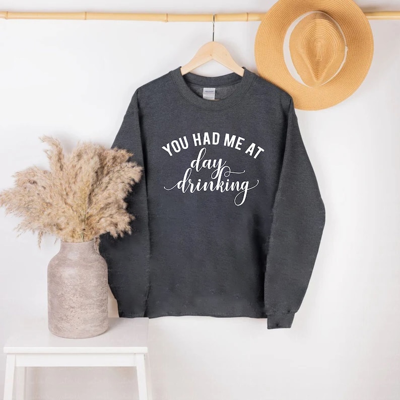 K&ouml;nnte beinhalten: Dunkelgraues Sweatshirt mit dem wei&szlig;en Text "YOU HAD ME AT day drinking" in einer Schreibschrift. Das Sweatshirt h&auml;ngt an einem Holzkleiderb&uuml;gel. Ein Strohhut und eine dekorative Vase befinden sich im Hintergrund.