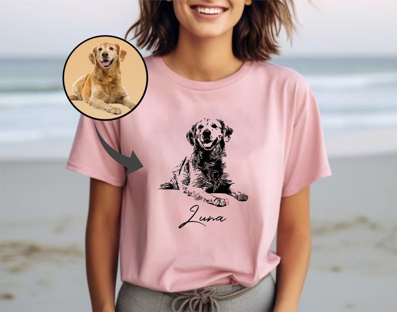 May include: A pink t-shirt featuring a black and white illustration of a dog. The dog is lying down and the text "Luna" is printed below the dog.