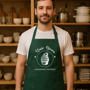 May include: A dark green apron with white text reading "Claire Brown Handmade Pastries". The apron has a cupcake design and two pockets. The person is wearing a white t-shirt in a kitchen setting.