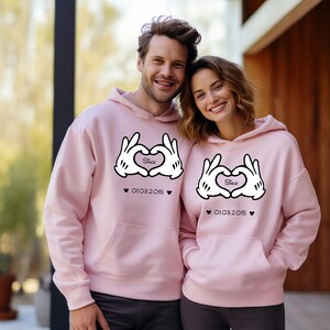 May include: Matching light pink hoodies with a heart-shaped hand design and the word "Since" inside the heart. The date "01.03.2015" is printed below the design. The hoodies have a front pocket and a hood.