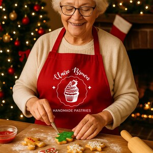 May include: A person wearing a red apron with the text "Claire Brown Handmade Pastries" decorates Christmas biscuits. The biscuits are on a wooden board and include gingerbread men, Christmas trees, and snowflakes. A rolling pin is visible.