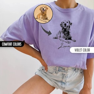 K&ouml;nnte beinhalten: Ein lila T-Shirt mit einer schwarz-wei&szlig;en Illustration eines Hundes und dem Namen "Luna" darauf gedruckt. Das Shirt wird von einer Person in Jeans-Shorts getragen.
