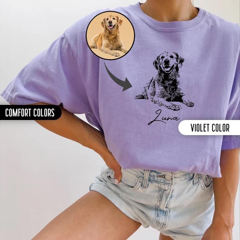 K&ouml;nnte beinhalten: Ein lila T-Shirt mit einer schwarz-wei&szlig;en Illustration eines Hundes und dem Namen "Luna" darauf gedruckt. Das Shirt wird von einer Person in Jeans-Shorts getragen.