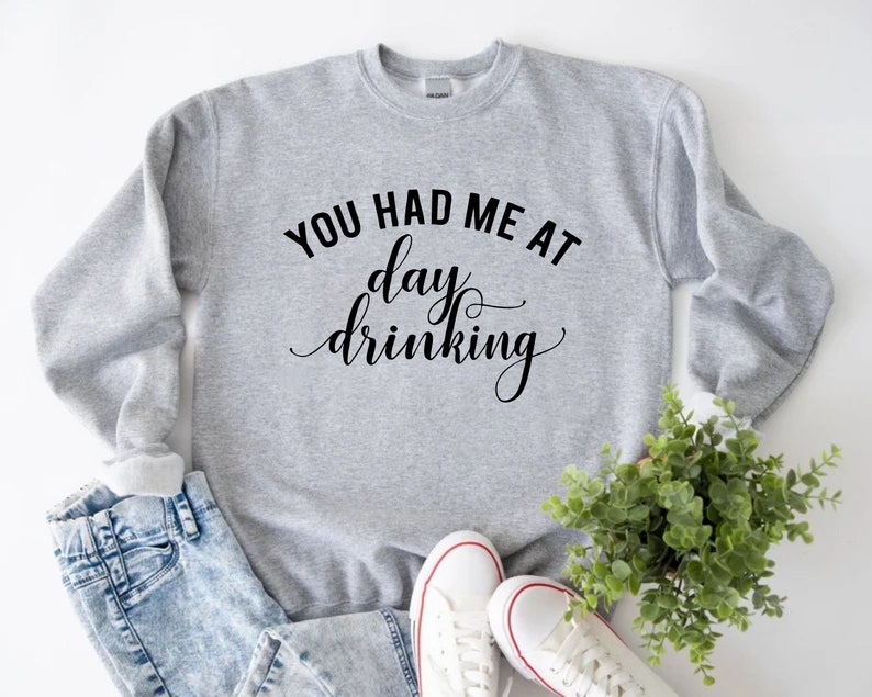 K&ouml;nnte beinhalten: Hellgraues Sweatshirt mit dem schwarzen Schriftzug "YOU HAD ME AT day drinking". Dazu eine hellblaue Jeans und wei&szlig;e Sneaker mit roten Akzenten. Eine kleine gr&uuml;ne Pflanze ist ebenfalls im Bild zu sehen.