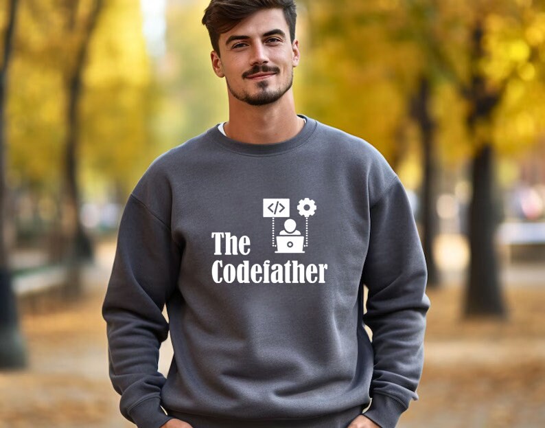 May include: A dark grey sweatshirt with a white graphic design. The graphic features a person sitting at a laptop with code symbols and a gear above them. The text "The Codefather" is below the graphic.