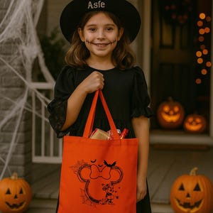 Puede incluir: Un bolso de tela de color naranja brillante con un estampado gr&aacute;fico negro de una silueta de orejas de rat&oacute;n con un lazo, un peque&ntilde;o murci&eacute;lago, una telara&ntilde;a y un peque&ntilde;o personaje de rat&oacute;n. El bolso tiene dos asas de tela naranja a juego.