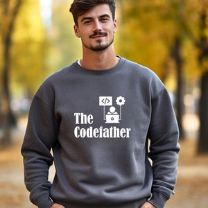 May include: A dark grey sweatshirt with a white graphic design. The graphic features a person sitting at a laptop with code symbols and a gear above them. The text "The Codefather" is below the graphic.