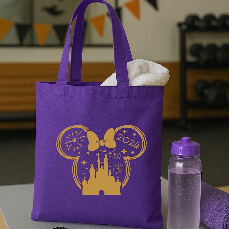 Puede incluir: Un bolso tote morado con un gr&aacute;fico dorado de un castillo, fuegos artificiales y un lazo con forma de orejas de Mickey Mouse. El texto "2020" est&aacute; en la oreja derecha. Una toalla blanca est&aacute; dentro de la bolsa. Una botella de agua morada y una esterilla de yoga enrollada.