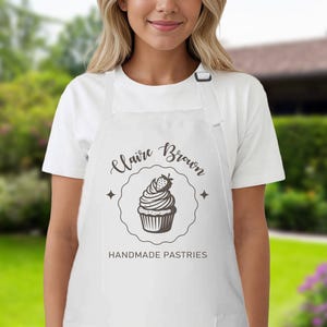 May include: White apron featuring a black cupcake design and the text "Claire Brown HANDMADE PASTRIES". The apron has a neck strap and two pockets. The design includes a cupcake with a strawberry.