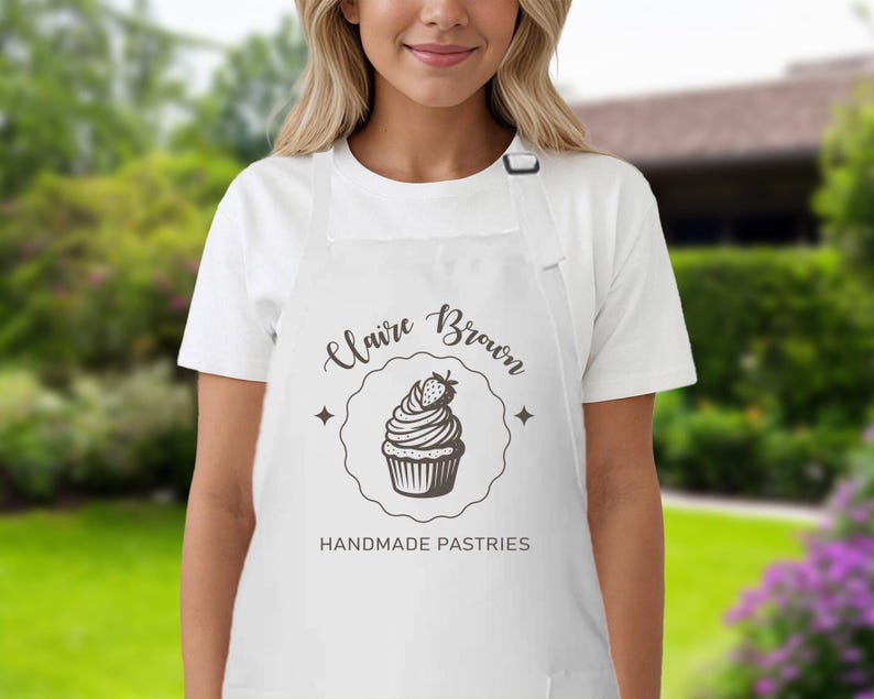 May include: White apron featuring a black cupcake design and the text "Claire Brown HANDMADE PASTRIES". The apron has a neck strap and two pockets. The design includes a cupcake with a strawberry.