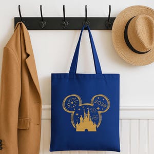 Puede incluir: Un bolso tote azul marino con un dise&ntilde;o dorado de orejas de Mickey Mouse. El dise&ntilde;o incluye un castillo, fuegos artificiales, estrellas y el a&ntilde;o 2020. La bolsa est&aacute; colgada de un gancho negro, junto a un abrigo color canela y un sombrero de paja.