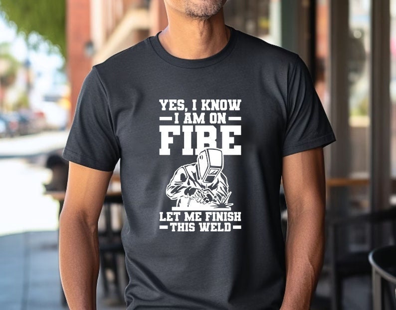 May include: A black t-shirt with white text that reads "Yes, I know - I am on fire - Let me finish this weld."  A white silhouette of a welder is shown welding.