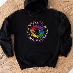 Pu&ograve; includere: Felpa con cappuccio nera con un simbolo di pace arcobaleno e un design a girasole. Il testo recita "Fai attenzione a chi odi, potrebbe essere qualcuno che ami."