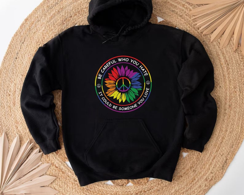 Pu&ograve; includere: Felpa con cappuccio nera con un simbolo di pace arcobaleno e un design a girasole. Il testo recita "Fai attenzione a chi odi, potrebbe essere qualcuno che ami."