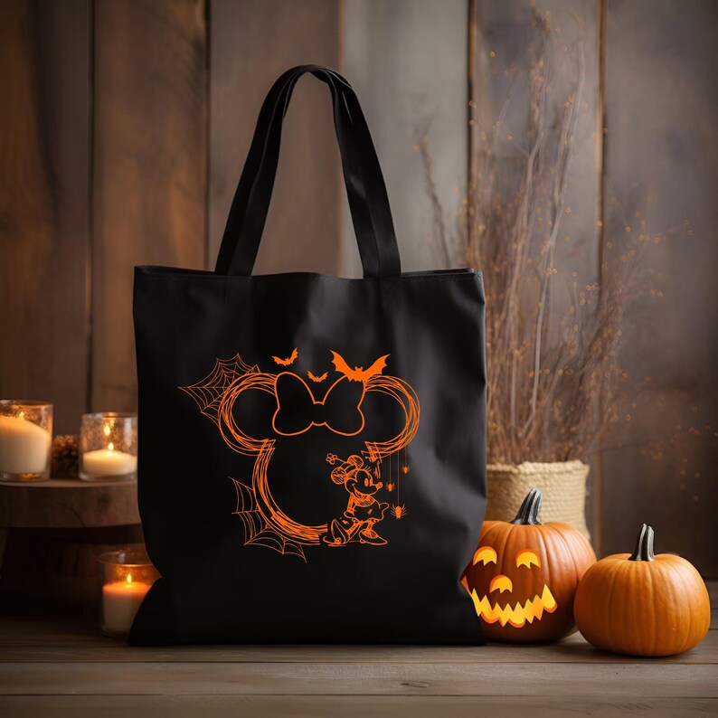 Puede incluir: Un bolso tote negro con un estampado gr&aacute;fico de color naranja brillante que muestra una silueta de orejas de rat&oacute;n con un lazo, un peque&ntilde;o personaje de rat&oacute;n, telara&ntilde;as y murci&eacute;lagos. El bolso cuenta con dos asas negras resistentes.