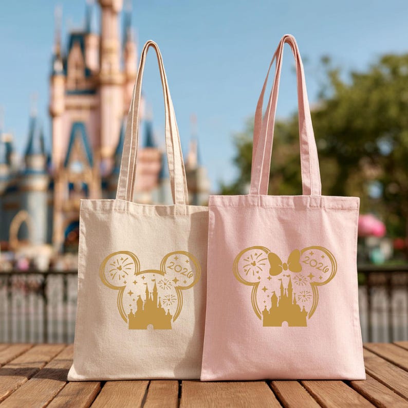 Puede incluir: Dos bolsas de lona, una beige y otra rosa, con dise&ntilde;os dorados de la cabeza de Mickey Mouse. El dise&ntilde;o incluye un castillo, fuegos artificiales y el a&ntilde;o 2026. Las bolsas tienen asas largas y est&aacute;n sobre una superficie de madera.