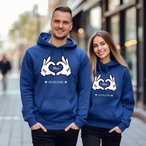 May include: Two matching blue hoodies with white Mickey Mouse hand heart design and the word "Since" inside the heart. The date "01.03.2015" is printed below the heart. The hoodies have a front pocket and a hood.