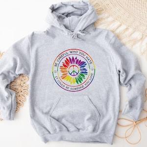 Pu&ograve; includere: Felpa con cappuccio grigia con un simbolo di pace arcobaleno e il testo "Fai attenzione a chi odi, potrebbe essere qualcuno che ami."