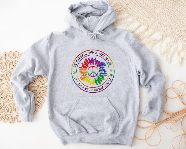 Pu&ograve; includere: Felpa con cappuccio grigia con un simbolo di pace arcobaleno e il testo "Fai attenzione a chi odi, potrebbe essere qualcuno che ami."