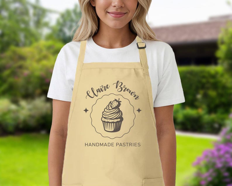 May include: A beige apron featuring the name "Claire Brown" and the words "Handmade Pastries" in black script. A cupcake illustration with a strawberry is in the center. The apron has a pocket and an adjustable neck strap.