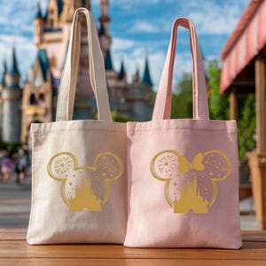 Puede incluir: Dos bolsas de mano de lona, una beige y otra rosa, con dise&ntilde;os dorados de la cabeza de Mickey Mouse. El dise&ntilde;o incluye una silueta de castillo, fuegos artificiales y el a&ntilde;o "2020". Ambas bolsas tienen asas a juego.