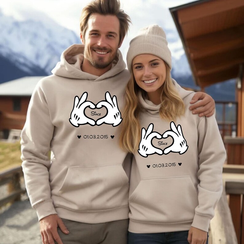 May include: Beige hoodies with a Mickey Mouse hand heart design. The design includes the word "Since" inside the heart, and the date 01.03.2015 below the heart. The hoodies have a front pocket and a hood.