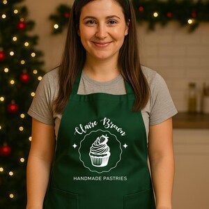May include: A woman wearing a green apron with the text "Claire Brown Handmade Pastries" and a reindeer antler headband. A selection of decorated Christmas biscuits are on a baking sheet in front of her.