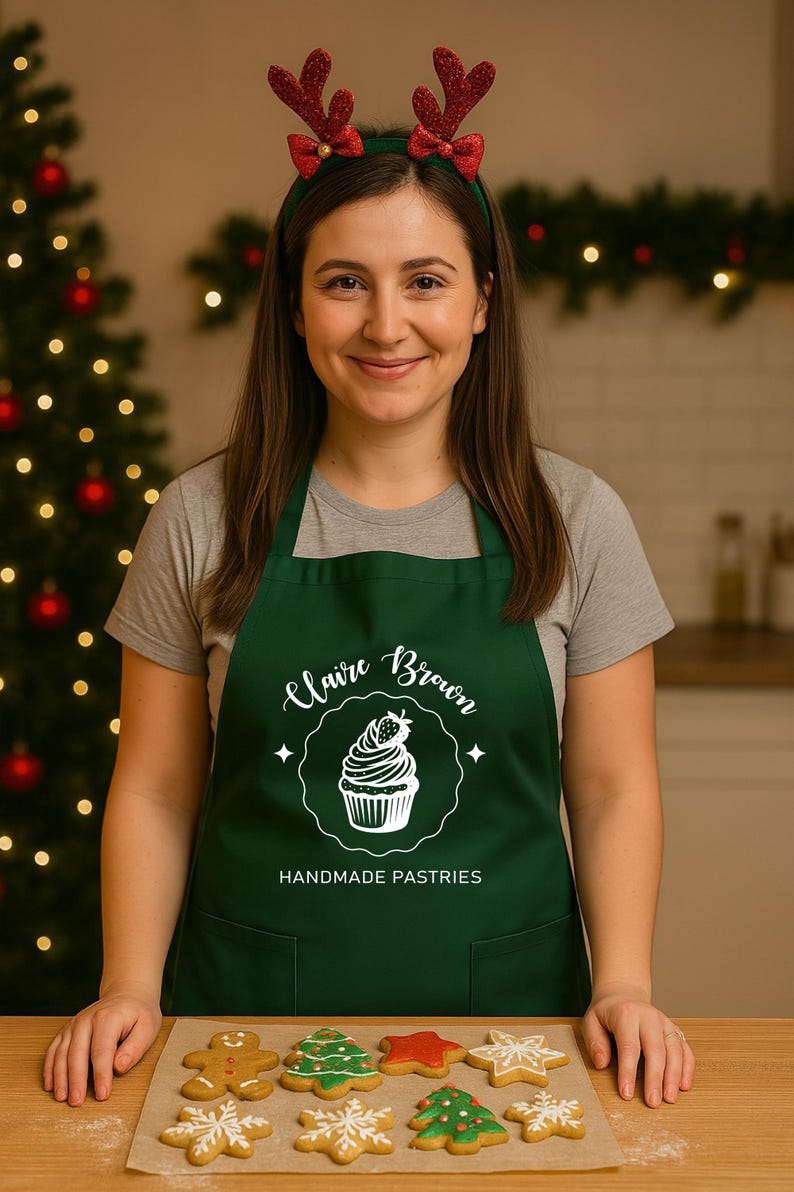 May include: A woman wearing a green apron with the text "Claire Brown Handmade Pastries" and a reindeer antler headband. A selection of decorated Christmas biscuits are on a baking sheet in front of her.