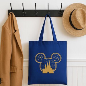 Puede incluir: Un bolso tote azul marino con un dise&ntilde;o dorado de orejas de Mickey Mouse. El dise&ntilde;o incluye un castillo, fuegos artificiales, estrellas y el a&ntilde;o 2020. La bolsa est&aacute; colgada de un gancho negro, junto a un abrigo color canela y un sombrero de paja.
