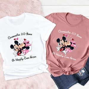 May include: Two t-shirts, one white and one dusty rose, featuring a cartoon image of Mickey and Minnie Mouse. The shirts have the text "Celebrating 20 Years Of Happily Ever After" above the image. The image shows Mickey and Minnie Mouse kissing.