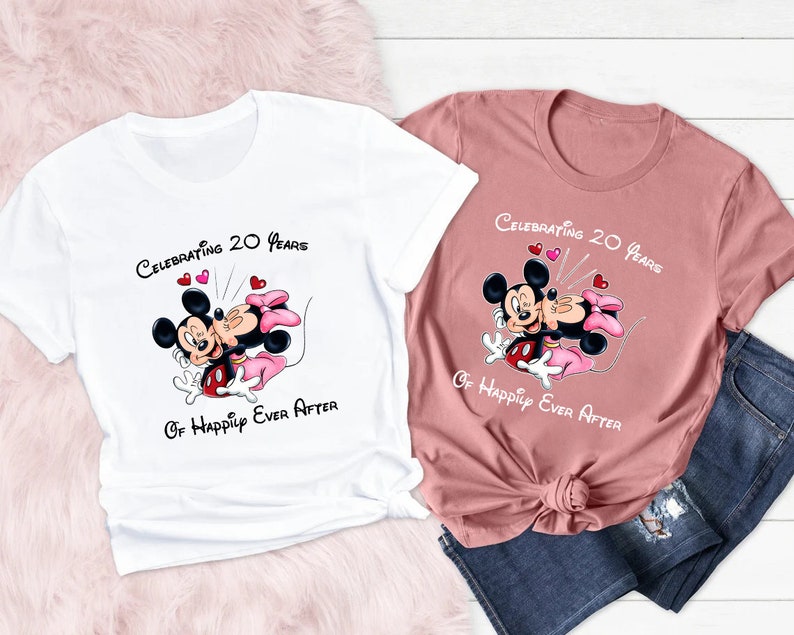 May include: Two t-shirts, one white and one dusty rose, featuring a cartoon image of Mickey and Minnie Mouse. The shirts have the text "Celebrating 20 Years Of Happily Ever After" above the image. The image shows Mickey and Minnie Mouse kissing.