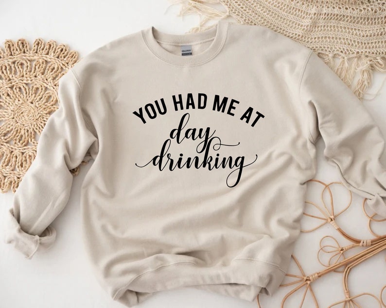 K&ouml;nnte beinhalten: Beiges Sweatshirt mit dem schwarzen Schriftzug "YOU HAD ME AT day drinking". Der Text ist in einer Mischung aus fetten und kursiven Schriftarten geschrieben. Das Sweatshirt hat einen Rundhalsausschnitt und lange &Auml;rmel.