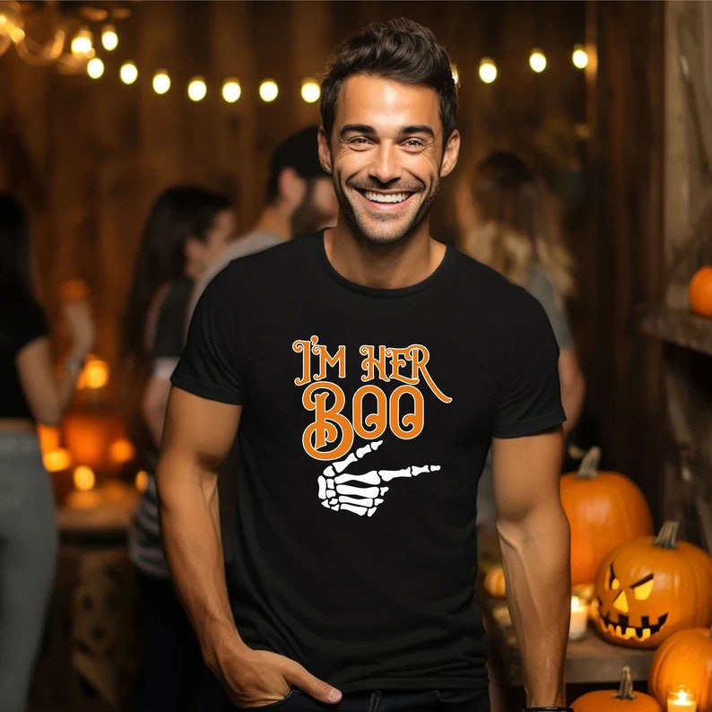 May include: A black t-shirt with the text "I'M HER BOO" in orange and white. The design includes a skeleton hand pointing to the right. The image is of a person smiling, with pumpkins in the background, suggesting a Halloween theme.