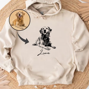May include: A cream-colored hoodie with a black and white illustration of a dog. The dog is lying down and the name "Luna" is written below the dog. There is a smaller image of a golden retriever in the top left corner.