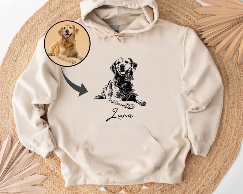 May include: A cream-colored hoodie with a black and white illustration of a dog. The dog is lying down and the name "Luna" is written below the dog. There is a smaller image of a golden retriever in the top left corner.
