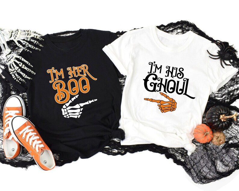 May include: Black and white t-shirts with Halloween-themed text and graphics. One shirt says "I'm Her Boo" with an orange and white skeleton hand pointing. The other shirt says "I'm His Ghoul" with a skeleton hand pointing.