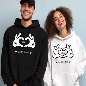 May include: Black and white matching hoodies. The black hoodie has a white graphic of gloved hands forming a heart, with the word "Since" inside the heart and the date "01.03.2015" below. The white hoodie has the same design.
