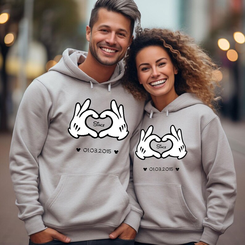 May include: Two light gray hoodies with a heart-shaped design made of white cartoon hands. The design includes the word "Since" inside the heart and the date "01.03.2015" below. The hoodies have a front pocket and a hood.