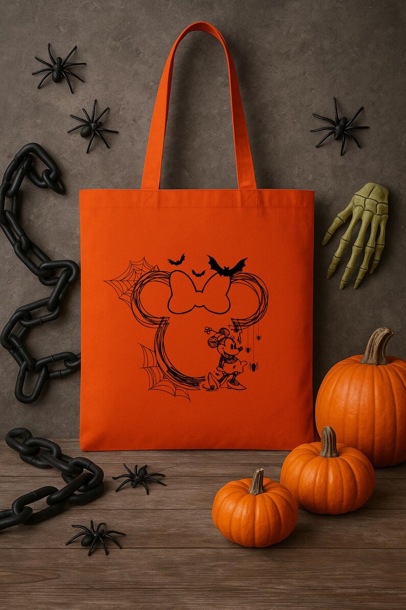 Puede incluir: Un bolso tote de color naranja brillante con un gr&aacute;fico de l&iacute;neas negras que muestra una silueta de orejas de rat&oacute;n con un lazo, una peque&ntilde;a figura de personaje, telara&ntilde;as, murci&eacute;lagos y ara&ntilde;as. El bolso tiene dos asas largas para el hombro.